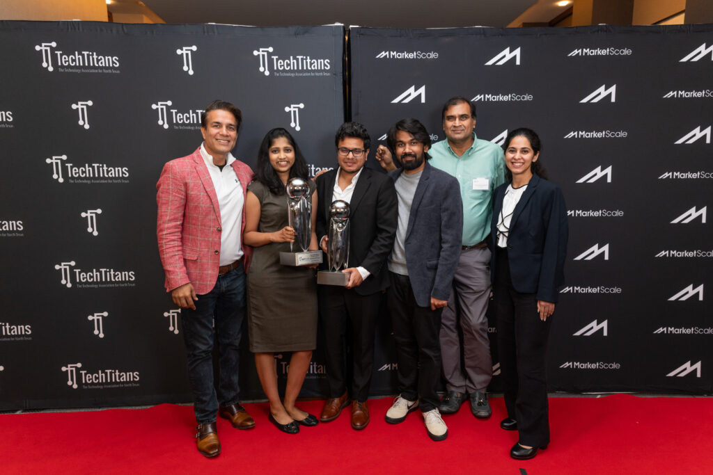 Group of seven people holding two award trophies on a red carpet in front of a Tech Titans backdrop, from the 2025 Tech Titans Awards.